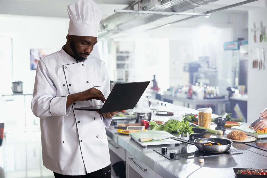 A male chef wearing a traditional white uniform and toque types on a laptop inside a brightly lit commercial kitchen. Stainless steel countertops around him are filled with fresh vegetables, pans, and food prep tools.