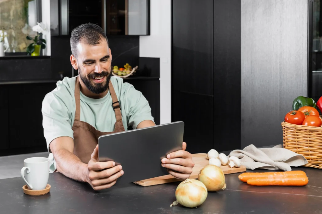 A smiling, bearded man wearing an apron holds a tablet while sitting at a modern kitchen counter. Fresh produce, including onions, carrots, mushrooms, and a basket of tomatoes, is spread across the dark surface alongside a coffee mug.