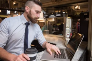 A bearded man dressed in a light blue button-down shirt and tie sits at a cafe counter while typing on a laptop. He holds a coffee cup with a lid as he intently focuses on his screen.