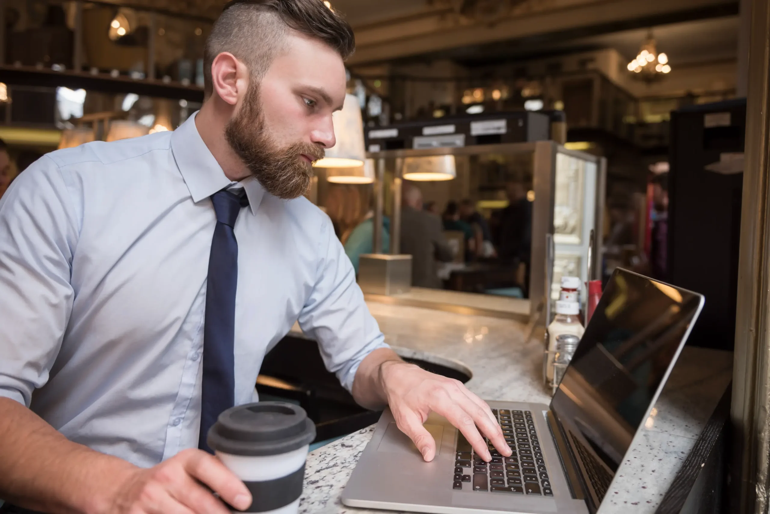 A bearded man dressed in a light blue button-down shirt and tie sits at a cafe counter while typing on a laptop. He holds a coffee cup with a lid as he intently focuses on his screen.