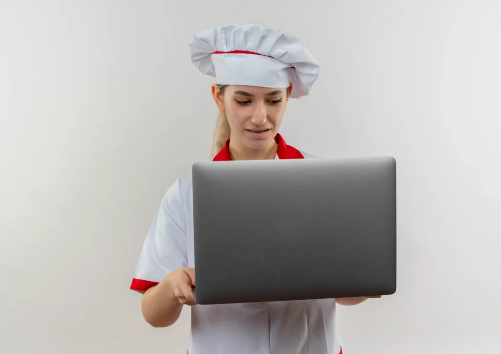 A young female chef in a white and red uniform holds an open gray laptop. She looks at the screen with an impressed expression against a plain white background.