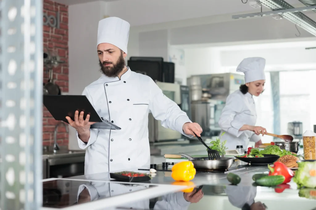 A bearded male chef holds a laptop in one hand while using a spatula to stir food in a skillet on a stovetop. In the background, another chef works at a counter surrounded by fresh ingredients in a modern commercial kitchen.
