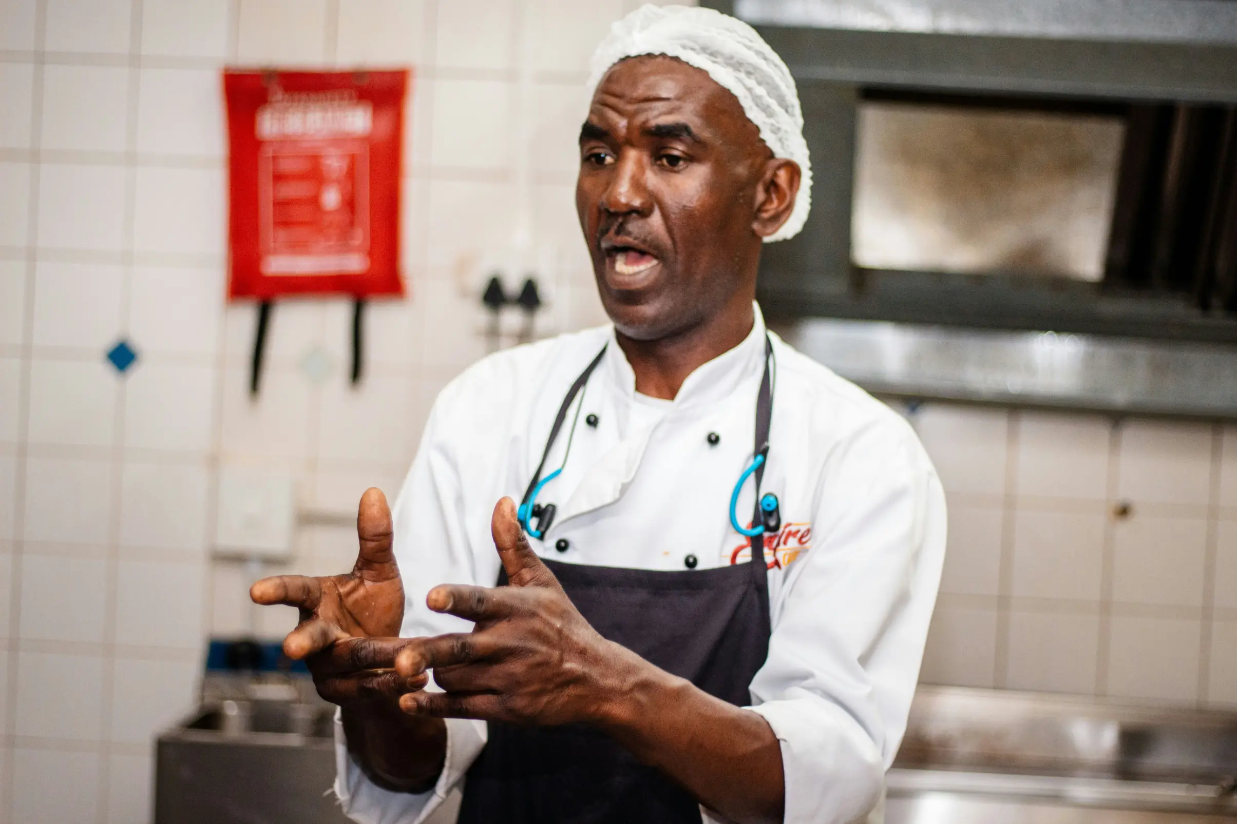 A male chef wearing a white hairnet, white double-breasted uniform, and black apron gestures with his hands while speaking. He stands in a commercial kitchen with white tiled walls and stainless steel equipment in the background.