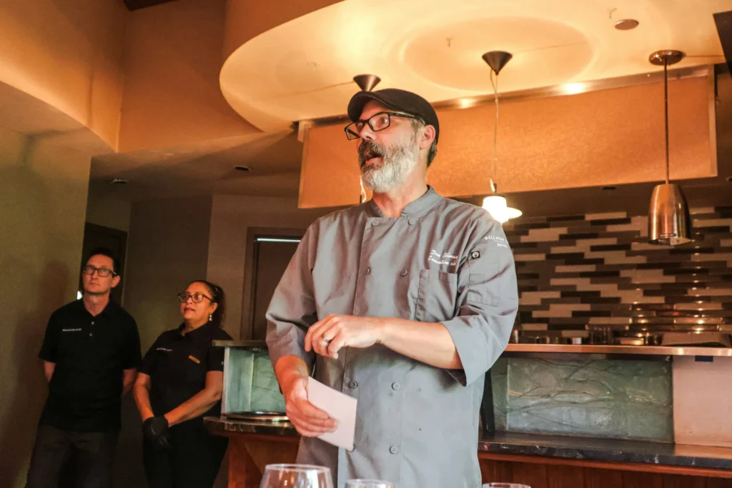 A chef with glasses, a grey beard, and a flat cap stands in a restaurant wearing a grey chef jacket while holding a card and addressing an audience. Two staff members in black shirts stand attentively in the background.