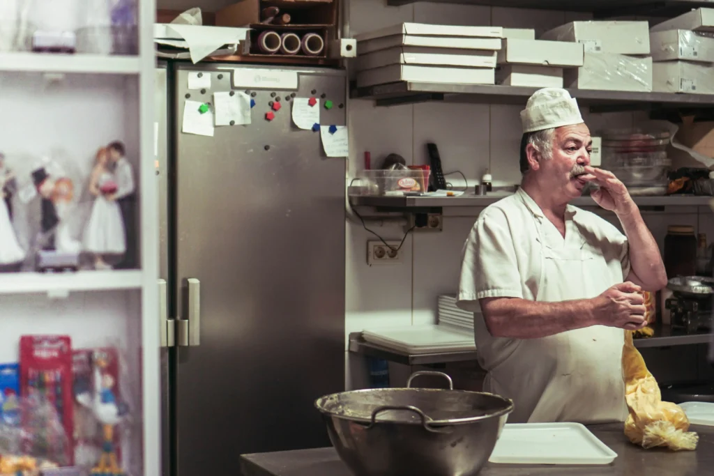 A baker in a white uniform and chef's hat licks his fingers while holding a piping bag filled with icing or dough. He stands in a busy kitchen filled with stainless steel appliances, stacked boxes, and cake decorating supplies.